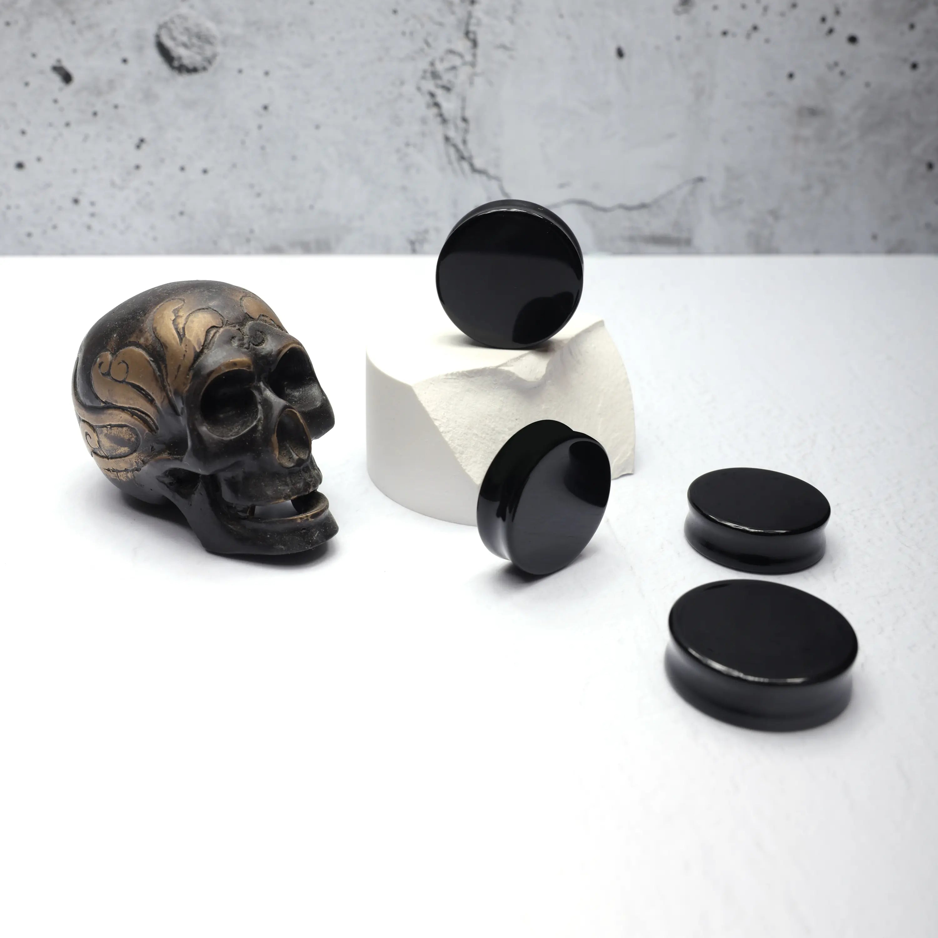 Black Obsidian Plugs - Product photo showing a collection of black obsidian plugs for large stretched ears spread out on a white foreground. A brass skull in the background gives the image attitude and fits with the overall branding